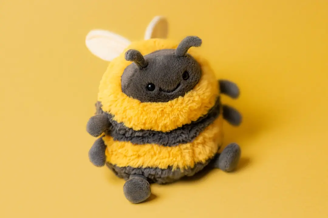 Jellycat Albee Bee plush toy with fluffy yellow and black stripes, smiling on a matching yellow background.